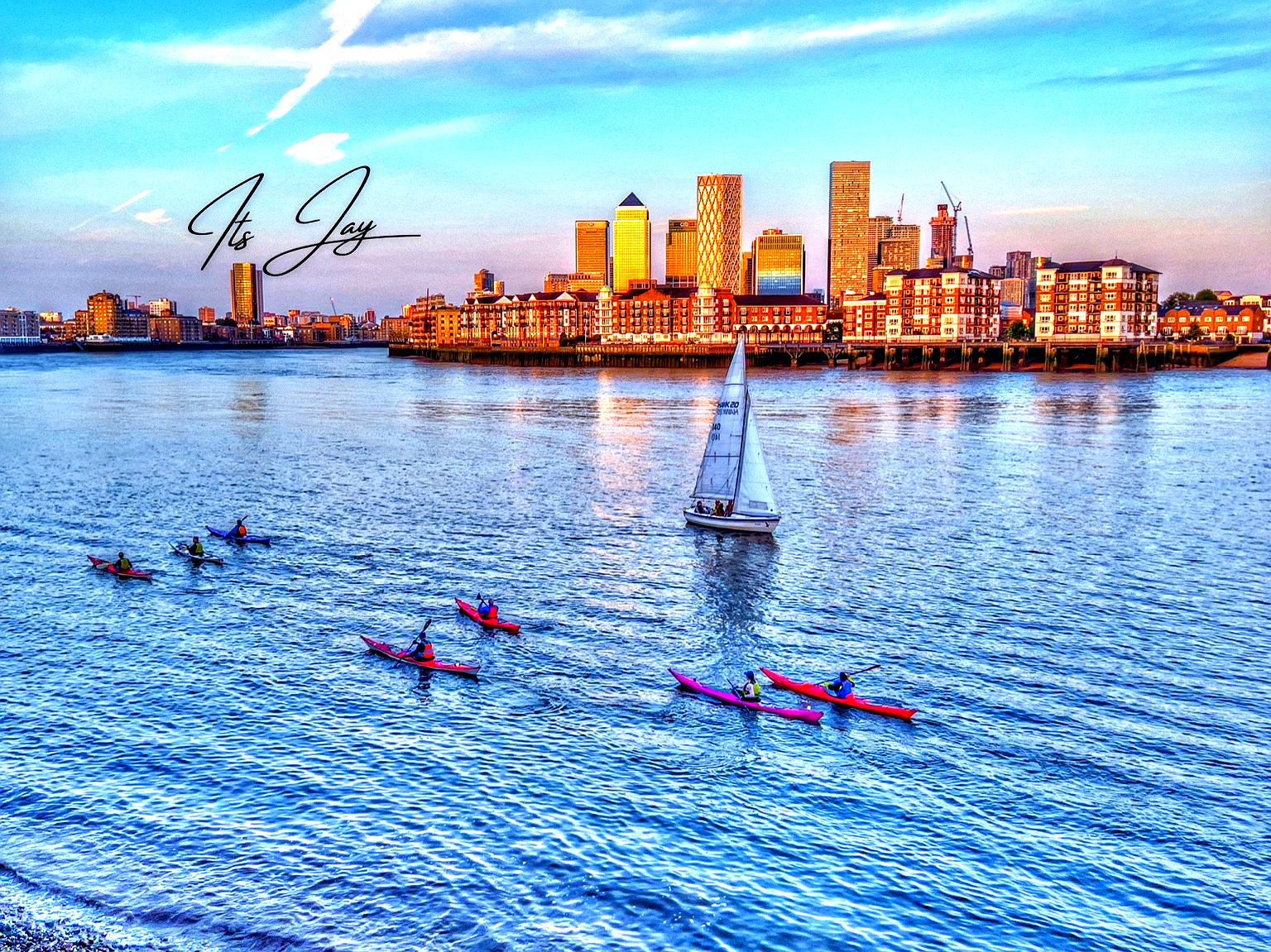 Colourful view of kayakers and a sailboat on the Thames with the Canary Wharf skyline in the background.